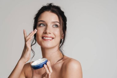 Kahverengi saçlı, gülümseyen beyaz bir kadın gri arka planda izole edilmiş bir kavanozdan yüz kremi sürüyor.