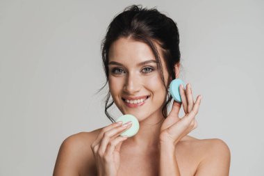 Kahverengi saçlı, gülümseyen beyaz kadın gri arka planda izole edilmiş güzellik blenderi kullanarak makyaj yapıyor.