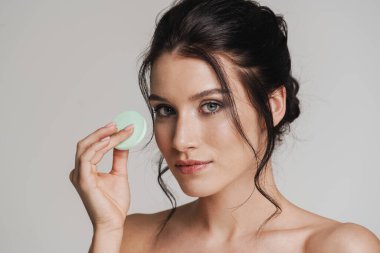 Kahverengi saçlı genç beyaz kadın gri arka planda izole edilmiş güzellik blenderi kullanarak makyaj yapıyor.