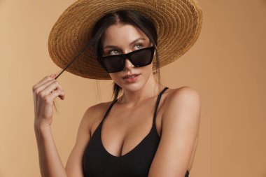 Hasır şapkalı genç esmer kadın izole edilmiş bej arka planda poz veriyor ve bakıyor.