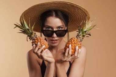 Hasır şapkalı genç esmer kadın ananaslı izole edilmiş bej arka planda poz veriyor.