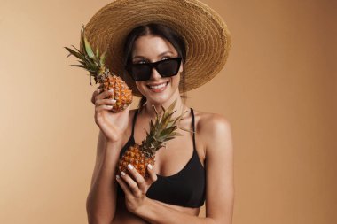 Hasır şapkalı genç esmer kadın ananaslı izole edilmiş bej arka planda poz verirken gülümsüyor.