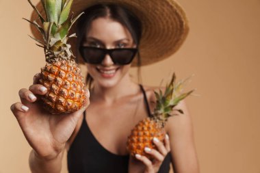 Hasır şapkalı genç esmer kadın ananaslı izole edilmiş bej arka planda poz verirken gülümsüyor.