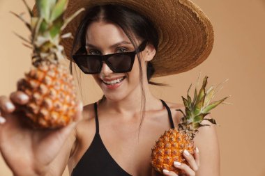 Hasır şapkalı genç esmer kadın ananaslı izole edilmiş bej arka planda poz verirken gülümsüyor.