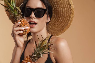 Hasır şapkalı genç esmer kadın ananaslı izole edilmiş bej arka planda poz veriyor.