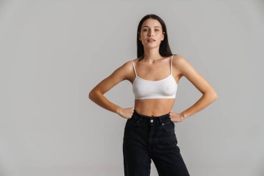 Gülümseyen beyaz kadın, uzun esmer saçlı, taraklı bluz giyen gri arka planda durup kameraya bakan.