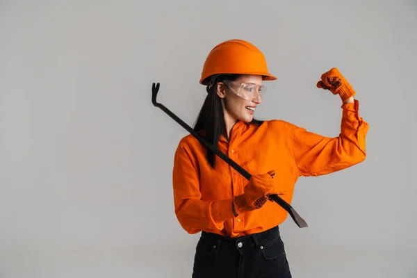 Young woman in helmet showing her bicep while posing with building tool isolated over grey background