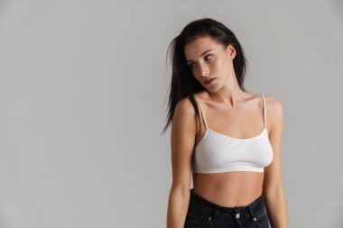 Genç esmer kadın, sütyen takıyor. Gri arka planda izole bir şekilde poz veriyor ve bakıyor.