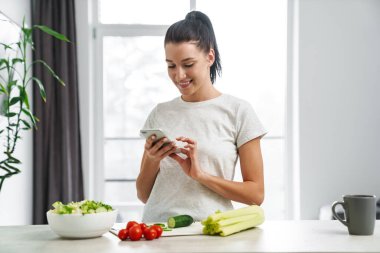 Avrupa 'dan gülümseyen kadın cep telefonu kullanıyor ve evde salata yapıyor.