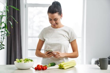 Avrupa 'dan gülümseyen kadın cep telefonu kullanıyor ve evde salata yapıyor.