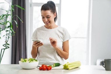 Avrupa 'dan gülümseyen kadın cep telefonu kullanıyor ve evde salata yapıyor.