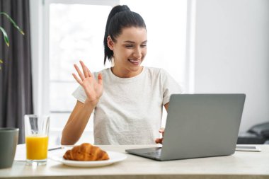 Avrupa 'dan gülümseyen bir kadın mutfakta kahvaltı ederken diz üstü bilgisayar kullanıyor.