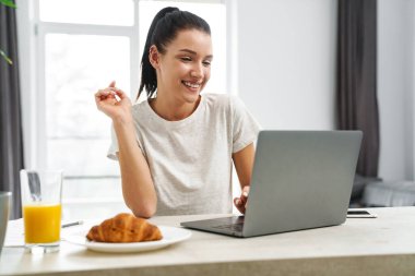 Avrupa 'dan gülümseyen bir kadın evde kahvaltı ederken dizüstü bilgisayar kullanıyor.