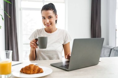Avrupa 'dan gülümseyen bir kadın evde kahve içerken dizüstü bilgisayar kullanıyor.