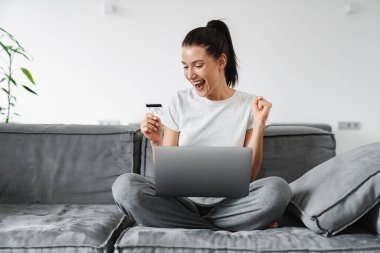 Heyecanlı kadın evde kredi kartı ve dizüstü bilgisayar kullanırken kazanan hareketi yapıyor