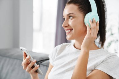 Evdeki koltukta otururken cep telefonu ve kulaklık kullanan gülümseyen kadın.