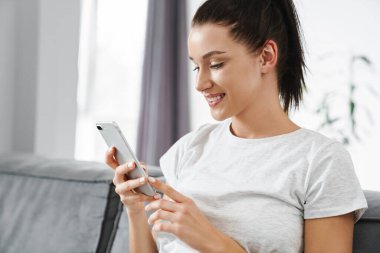 Avrupa 'dan gülümseyen bir kadın evdeki koltukta otururken cep telefonu kullanıyor.