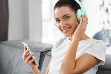 Evdeki koltukta otururken cep telefonu ve kulaklık kullanan gülümseyen kadın.