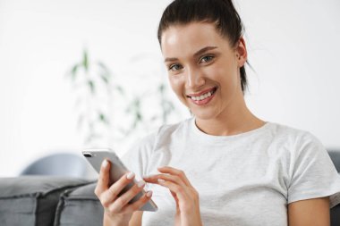 Avrupa 'dan gülümseyen bir kadın evdeki koltukta otururken cep telefonu kullanıyor.