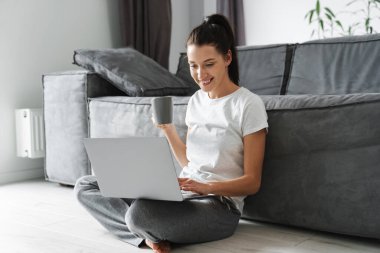 Gülümseyen kadın kahve içiyor ve evde otururken dizüstü bilgisayar kullanıyor.