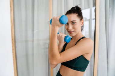 Mutlu esmer beyaz kadın oturma odasında dambılları kaldırıyor.