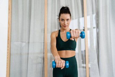 Mutlu esmer beyaz kadın oturma odasında dambılları kaldırıyor.