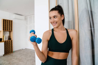 Mutlu esmer beyaz kadın oturma odasında dambılları kaldırıyor.