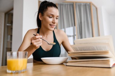 Beyaz esmer kadın evde kahvaltı ederken kitap okuyor.