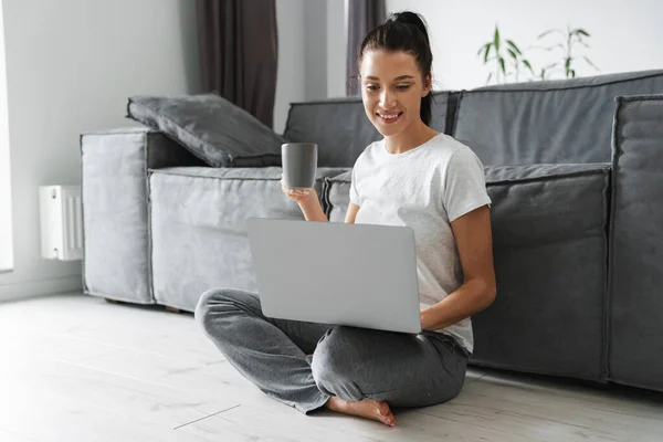 Lächelnde Frau Trinkt Kaffee Und Benutzt Laptop Während Sie Hause — Stockfoto