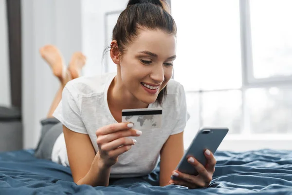 Smiling Woman Holding Credit Card Using Cellphone While Lying Bed — Stock Photo, Image