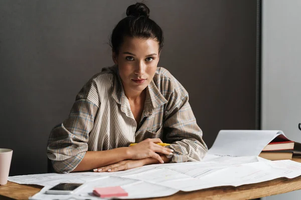 Young brunette woman working with drawings while sitting at desk in office