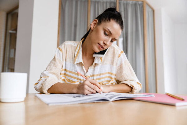 White brunette woman talking on cellphone while doing homework at home