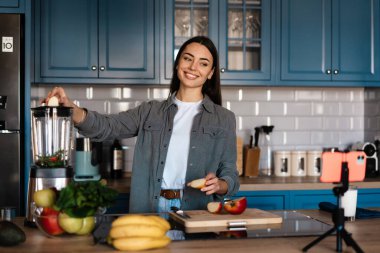 Gülümseyen kadın ev mutfağında cep telefonundan selfie çekerken smoothie yapıyor.