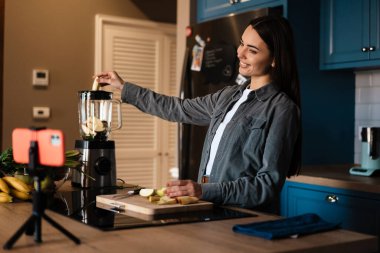 Gülümseyen kadın ev mutfağında cep telefonundan selfie çekerken smoothie yapıyor.