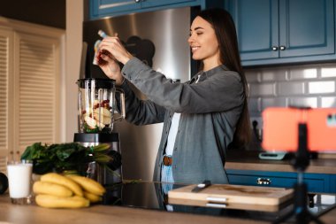 Gülümseyen kadın ev mutfağında cep telefonundan selfie çekerken smoothie yapıyor.