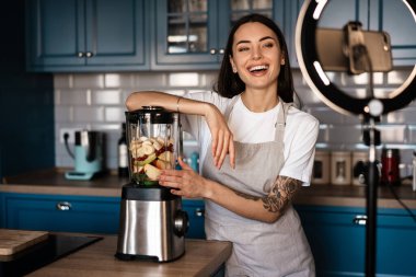 Ev mutfağında cep telefonundan selfie çekerken gülen kadın smoothie yapıyor.
