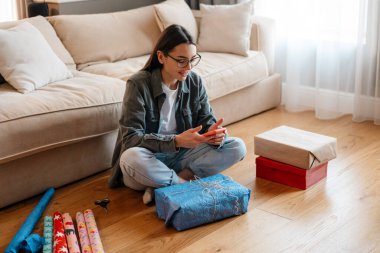Evde hediyeleri sararken cep telefonu kullanan beyaz bir kadın.