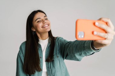 Beyaz arka planda izole edilmiş cep telefonuyla selfie çeken genç bir kadın.
