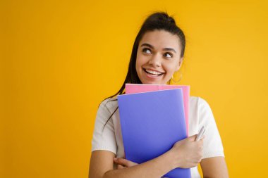 Sarı arka planda izole edilmiş egzersiz kitaplarıyla poz verirken gülümseyen genç esmer kadın.