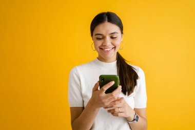 Sarı arka planda izole edilmiş gülümseyen ve cep telefonu kullanan genç esmer kadın.