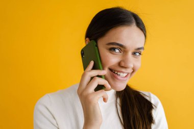 Sarı arka planda izole edilmiş, gülümseyen ve cep telefonuyla konuşan genç esmer kadın.