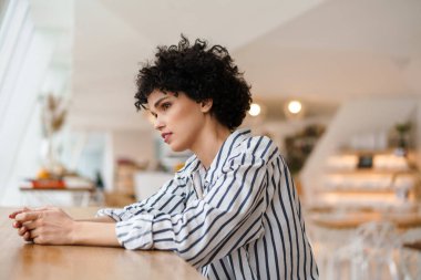 Kafede otururken pencereden dışarı bakan güzel, odaklanmış kıvırcık kadın.