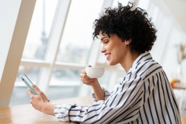 Güzel, gülümseyen kıvırcık kadın kafede kahve içerken cep telefonu kullanıyor.