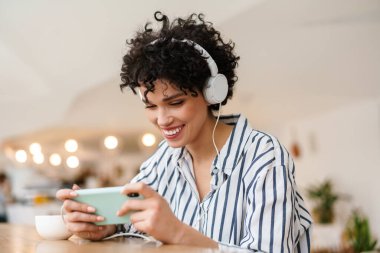 Kafede kahve içerken cep telefonu kullanan kulaklıklı hoş, neşeli bir kadın.