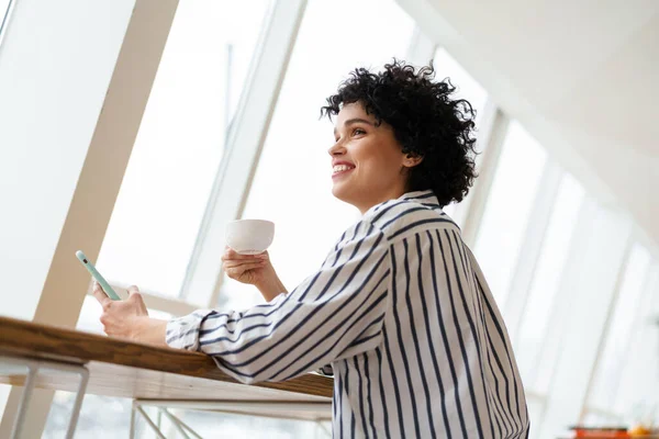 Hermosa Mujer Rizada Sonriente Usando Teléfono Móvil Mientras Bebe Café —  Fotos de Stock