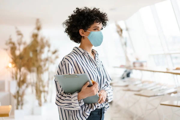 Beautiful Curly Nice Woman Face Mask Holding Clipboard While Standing — Stock Photo, Image