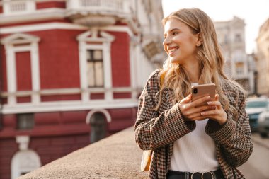 Gülümseyen genç sarışın kadın cep telefonu kullanıyor ve caddede yürüyor.