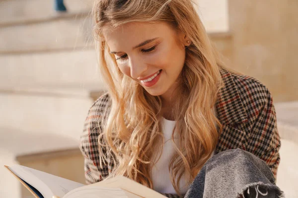 Улыбающаяся Молодая Блондинка Читающая Книжку Сидя Улице Закрывается — стоковое фото