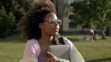 Kıvırcık saçlı, güneş gözlüğü takan Amerikalı bir kadın kampüsün dışında dizüstü bilgisayarını tutarken yürüyor.