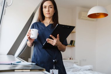 Mavi üniformalı, elinde kağıtlar ve elinde fincanla elinde tabletle odada pencerenin yanında dururken kameraya bakan kendine güvenen kadın.